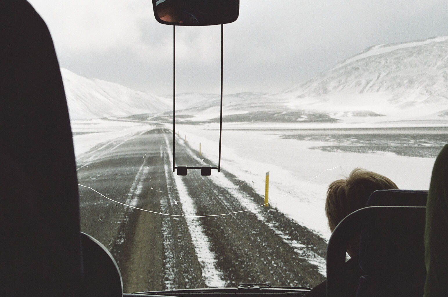 A snowy valley and partly snow-covered Route 1 with yellow road posts, seen from inside a car