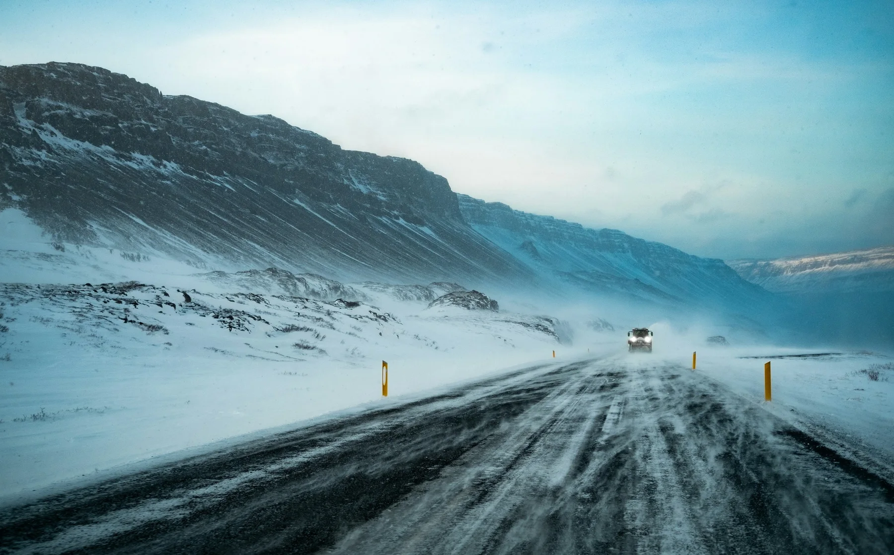 Snow-covered road in Iceland with blowing snow, yellow markers, and oncoming traffic