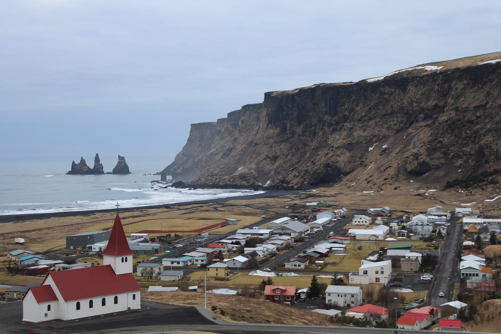 A real view of Vík village, its church, black-sand coast and cliffs; used to understand the spatial relationship between accommodation and the next day’s route, not to represent any hotel view or same-day road conditions